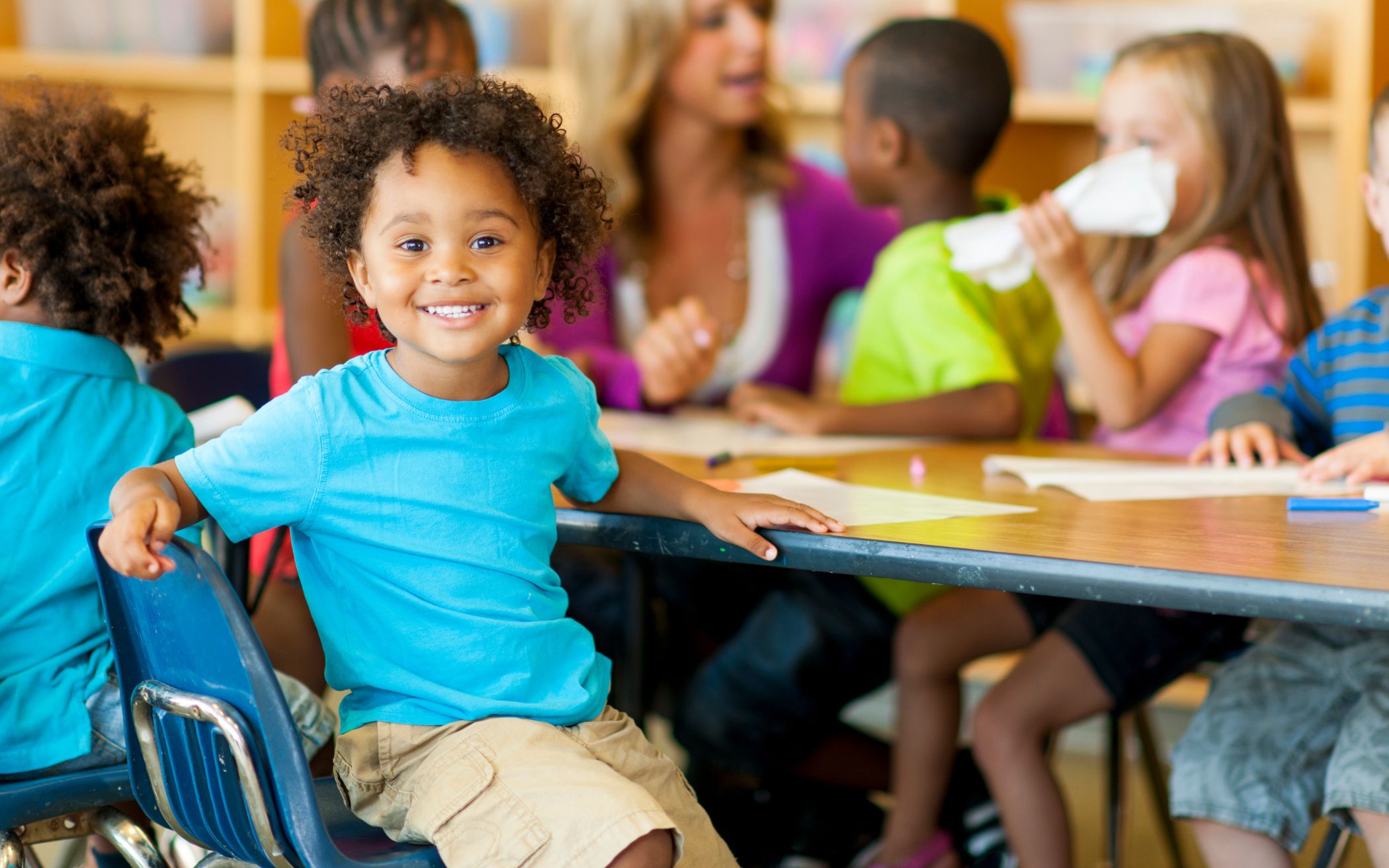Child in blue shirt sitting at a table smiling at the camera with other children and teacher in the background at preschool.