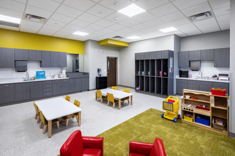 Toddler classroom with yellow rug and accents, tables and chairs, and storage cabinets.
