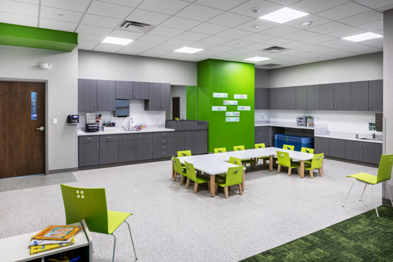 Toddler room with green accents and tables and chairs and storage cabinets.